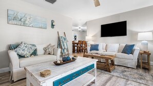 A living room with a white sofa, a coffee table with a sign, and a TV mounted on the wall.