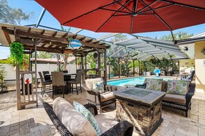 A covered outdoor area with a pool, wicker furniture, and a pergola with a red umbrella. - Twin Oaks Luxury Heated Pool and Caribbean Tiki Bar Near Indian Rocks Beach (Largo)
