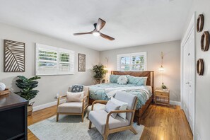A bedroom with a bed, two chairs, a ceiling fan, and artwork on the walls.