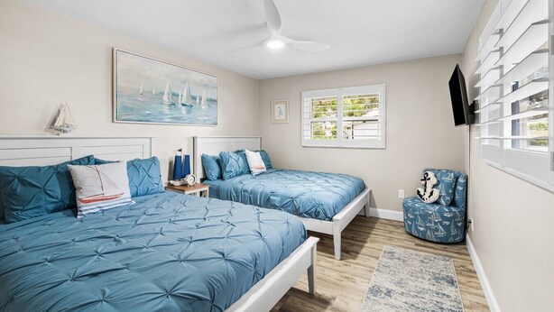 A bedroom with two beds, a ceiling fan, a painting, a window, and a TV.