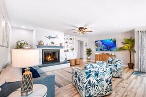 Una sala de estar con chimenea, ventilador de techo, televisor de pantalla plana y un gran ventanal con cortinas.