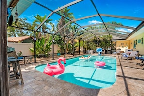 A covered outdoor swimming pool with two pink flamingo floats, surrounded by lounge chairs and a wooden deck.