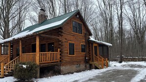 Una cabaña de troncos con porche y chimenea, rodeada de nieve y árboles.