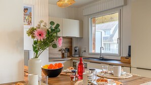 Een moderne keuken met witte kasten, een houten aanrechtblad en een groot raam. Links staat een vaas met bloemen, op het aanrecht een schaal met mandarijnen en een rode fles.