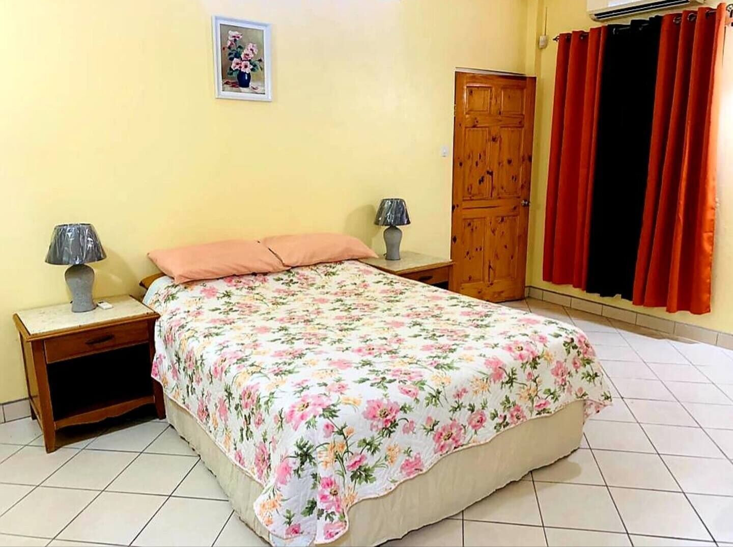 Un dormitorio con una cama, dos mesitas de noche con lámparas, un armario de madera y una ventana con cortinas rojas.