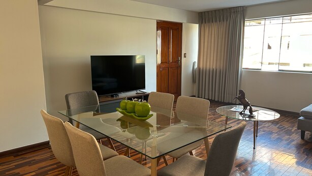 A modern living room with a glass dining table, six chairs, a flat-screen TV, and a decorative apple bowl.