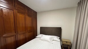 A hotel room with a large wooden wardrobe, a single bed with white bedding, a nightstand, and a curtain.