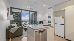 Una cocina moderna con una isla central, lavavajillas integrado y un refrigerador. Hay una zona de estar con sofá y una mesa de centro, y un amplio ventanal con vistas a la ciudad.