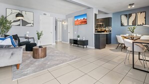 Una sala de estar moderna con un sofá gris, una mesa de centro y un comedor con mesa y sillas. Se observa una cocina con armarios oscuros y un microondas.