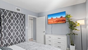 Un dormitorio con cama, cómoda, planta en maceta, televisor en la pared y puerta que da a otra habitación.