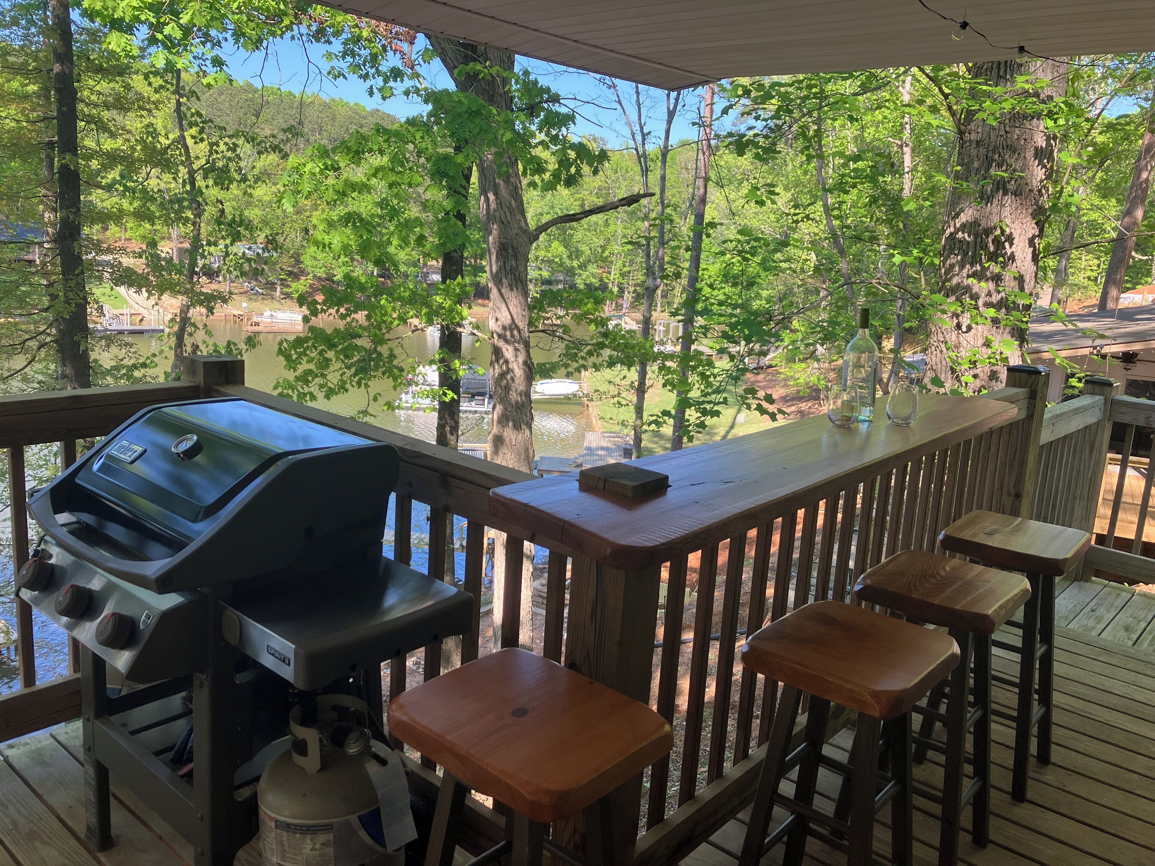 Una terraza techada con una parrilla, taburetes de madera y vistas a un lago y árboles.