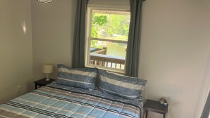 Ein Schlafzimmer mit einem Bett, einem Nachttisch, einer Lampe, einem Fenster mit Blick auf eine Terrasse und Grünflächen sowie einem kleinen Tisch mit Lampe.