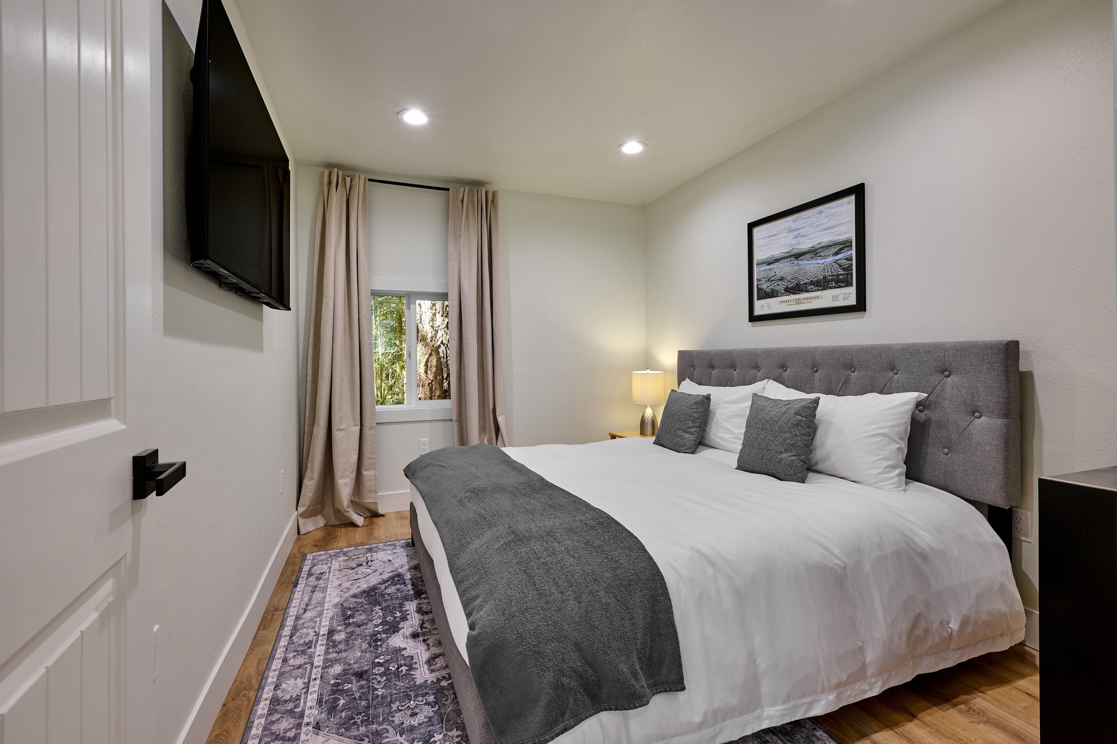 A bedroom with a bed, a TV mounted on the wall, a window with curtains, and a framed picture on the wall.