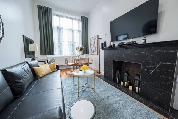 Een moderne woonkamer met een zwarte leren bank, een witte salontafel, een open haard en een grote flatscreen televisie aan de muur.