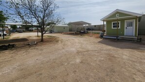 A small, single-story building with a green exterior and a white door, surrounded by a gravel driveway and a tree. - Tinyhouse with AC and laundry area. Near Ector County Coliseum (Odessa)