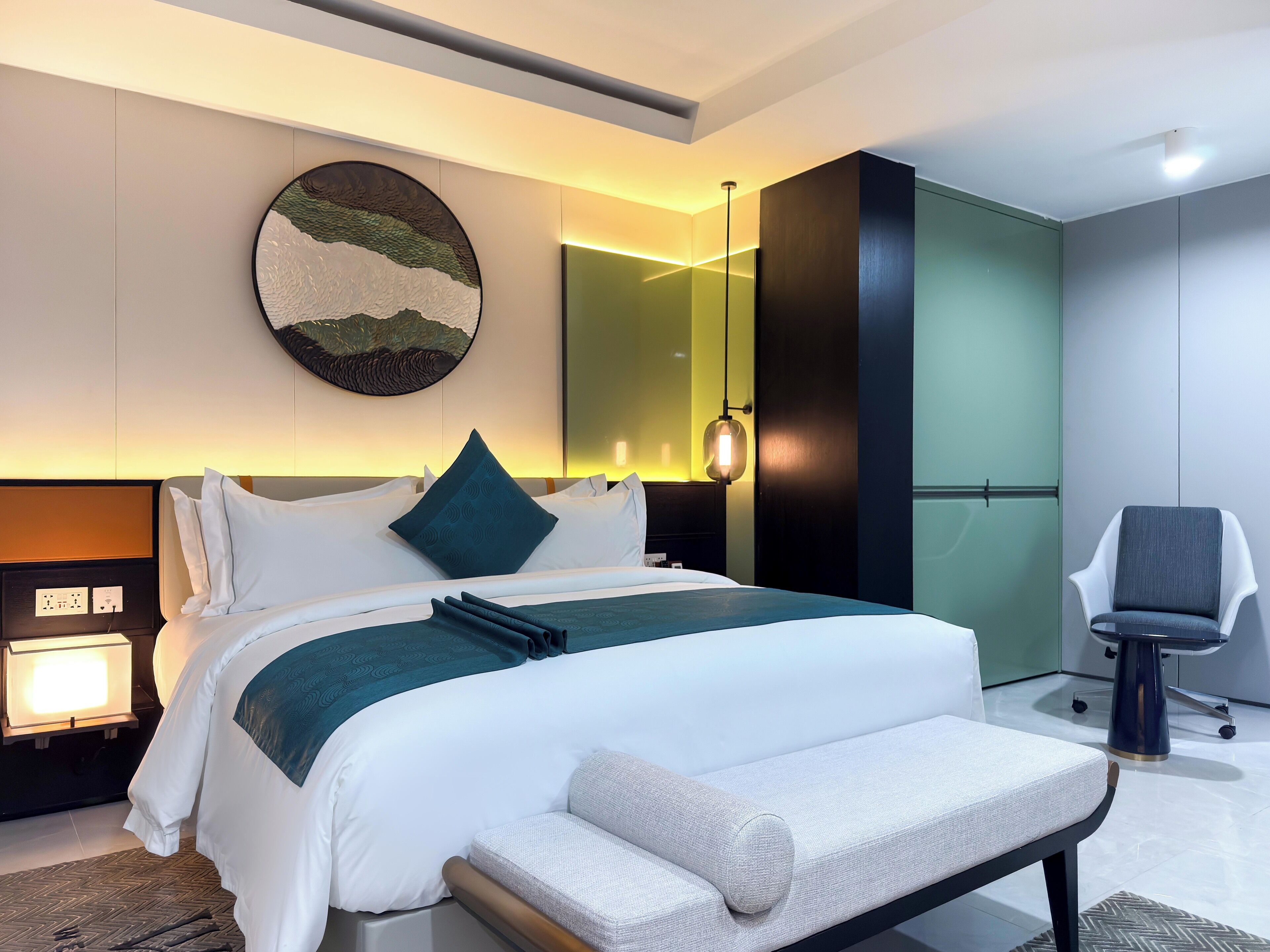 A modern hotel room with a neatly made bed, a large wall-mounted artwork, a bedside table with a lamp, and a chair.