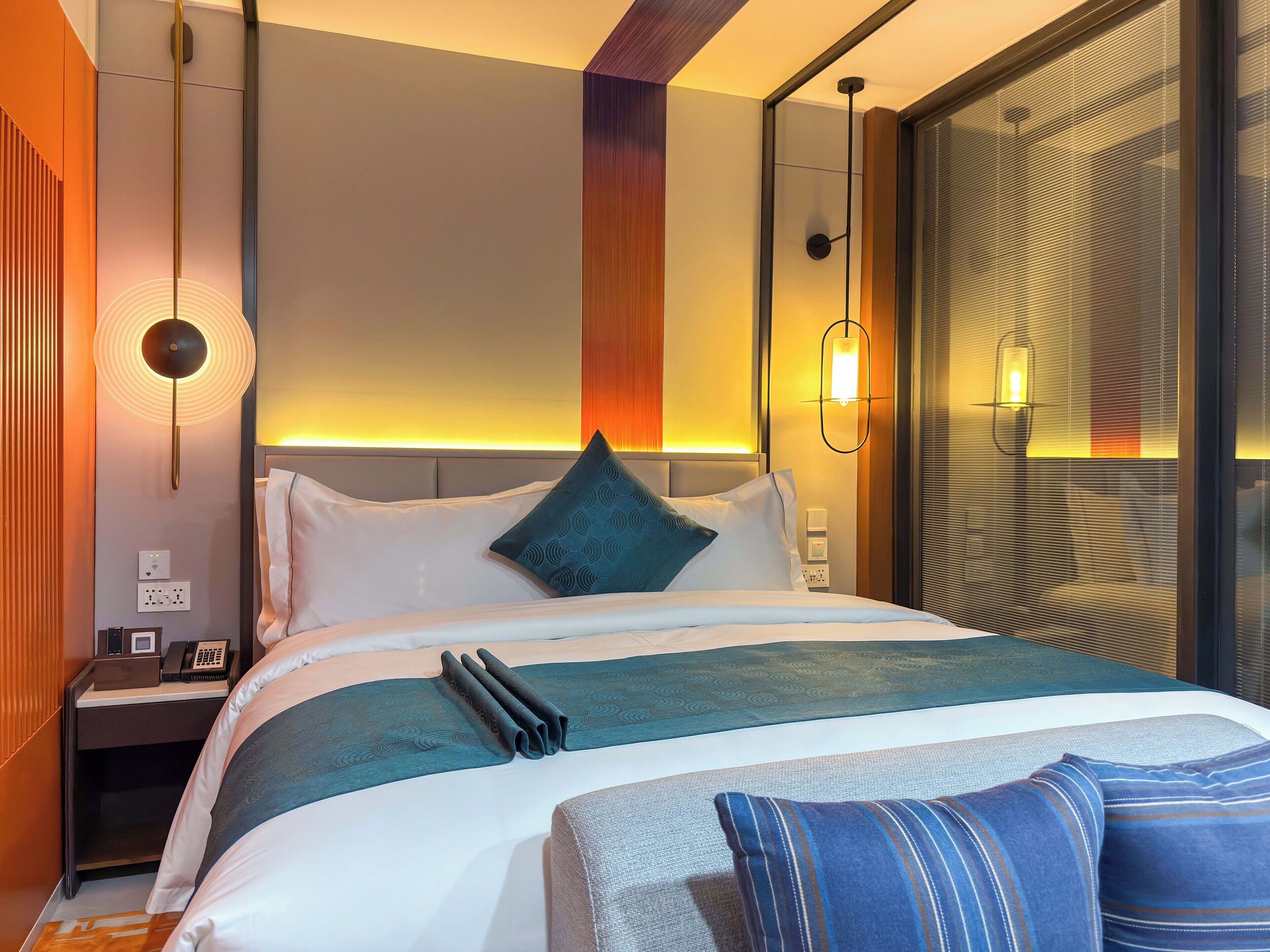 A modern hotel room with a neatly made bed, a bedside table with a phone, and a wall-mounted lamp.