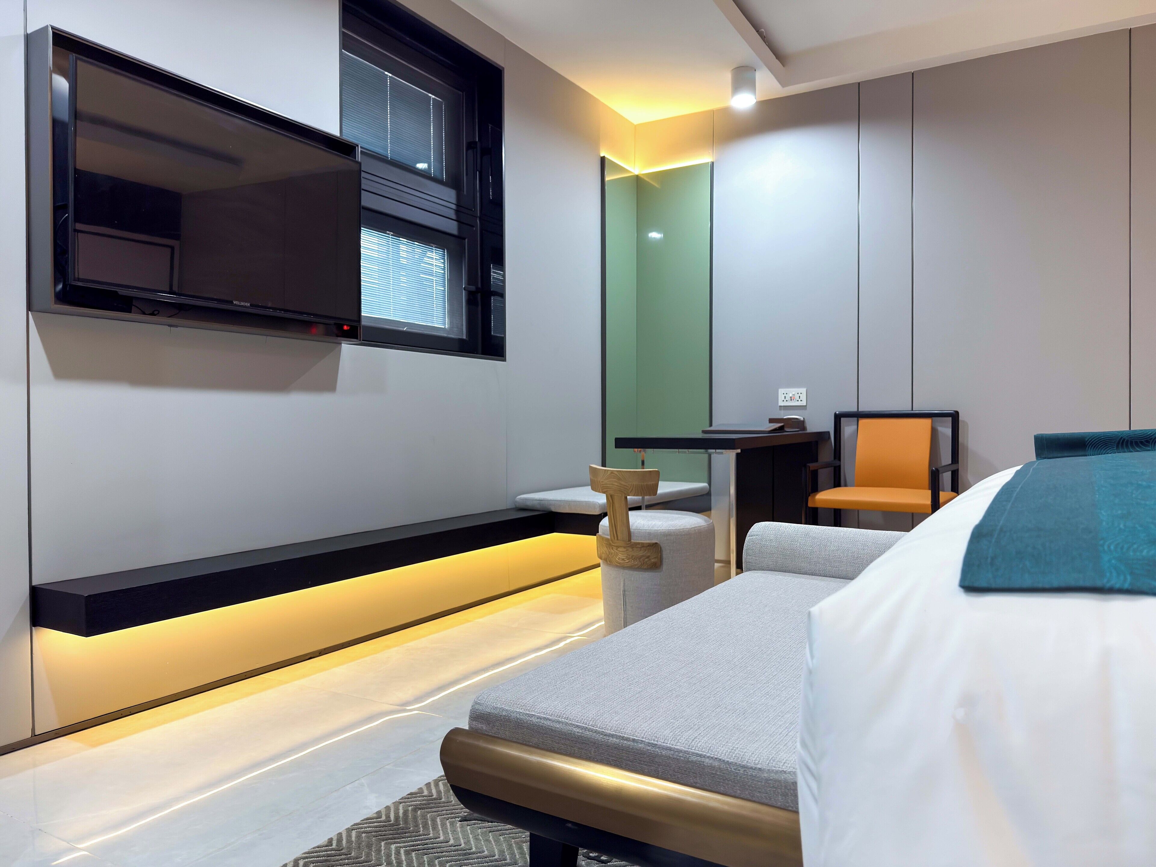 Una habitación de hotel moderna con televisión de pantalla plana montada en la pared, un escritorio pequeño con una silla y una cama con una manta azul.