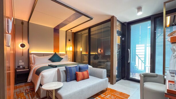 Ett modernt hotellrum med en stor säng, en soffa, ett litet bord och en stol. Det finns ett stort fönster med utsikt över staden.