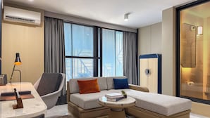 Ett modernt hotellrum med en bäddsoffa, ett skrivbord med en lampa, ett litet bord och ett stort fönster med gardiner.