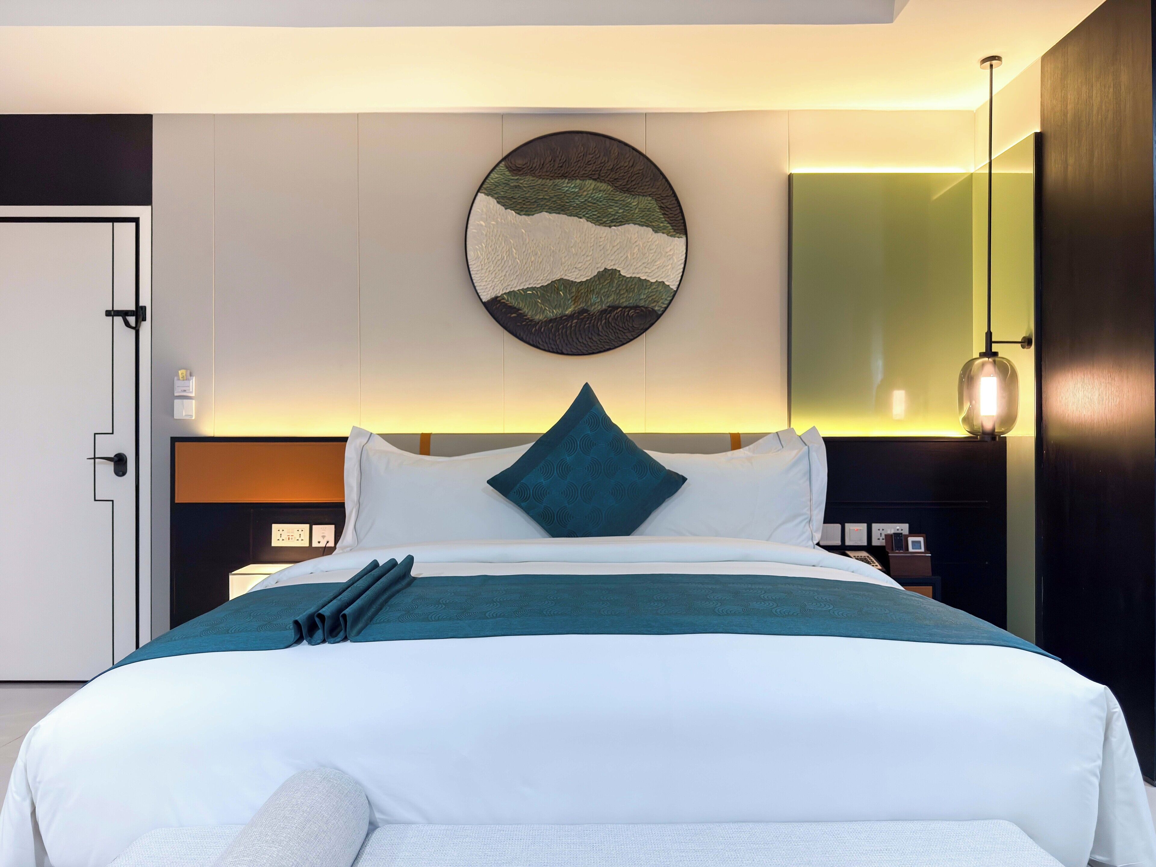 A modern hotel room with a neatly made bed, a round wall art piece, a large mirror, and a bedside table with a lamp.