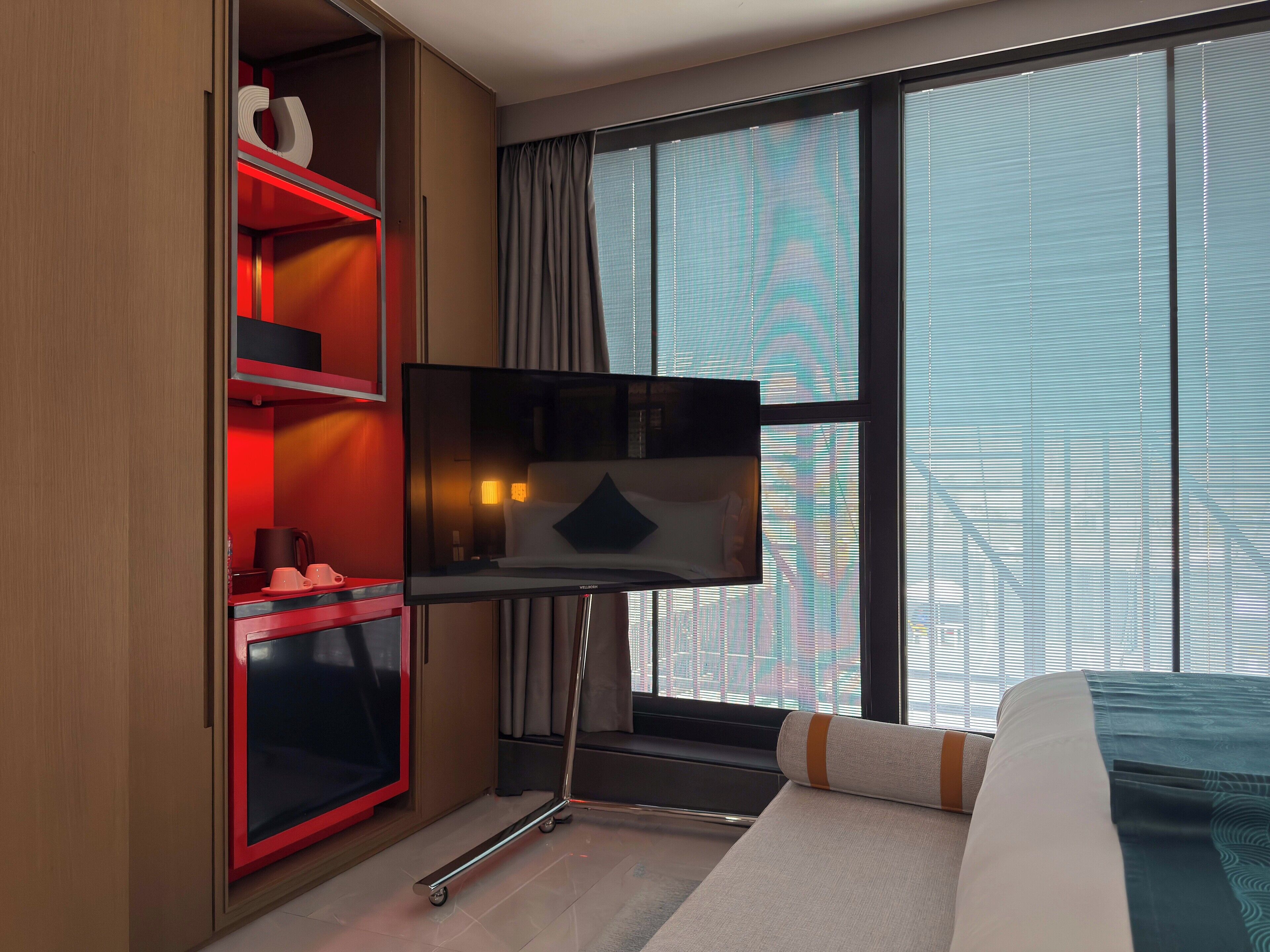 Ein Hotelzimmer mit Bett, Fernseher auf einem Schrank, Kleiderschrank und großem Fenster mit Jalousien.