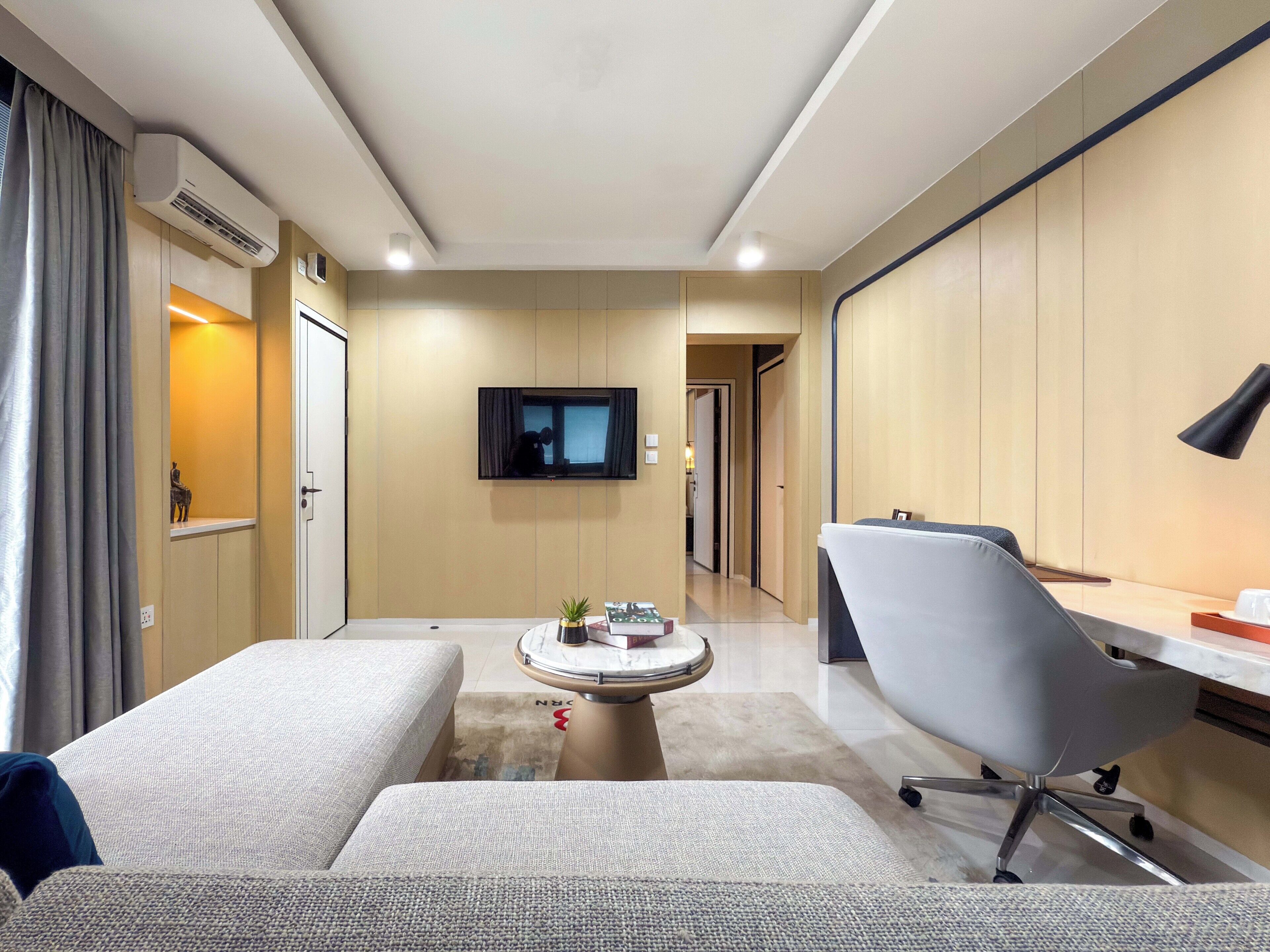 A modern hotel room with a sofa, a desk with a chair, a television mounted on the wall, and a small table with decorative items.