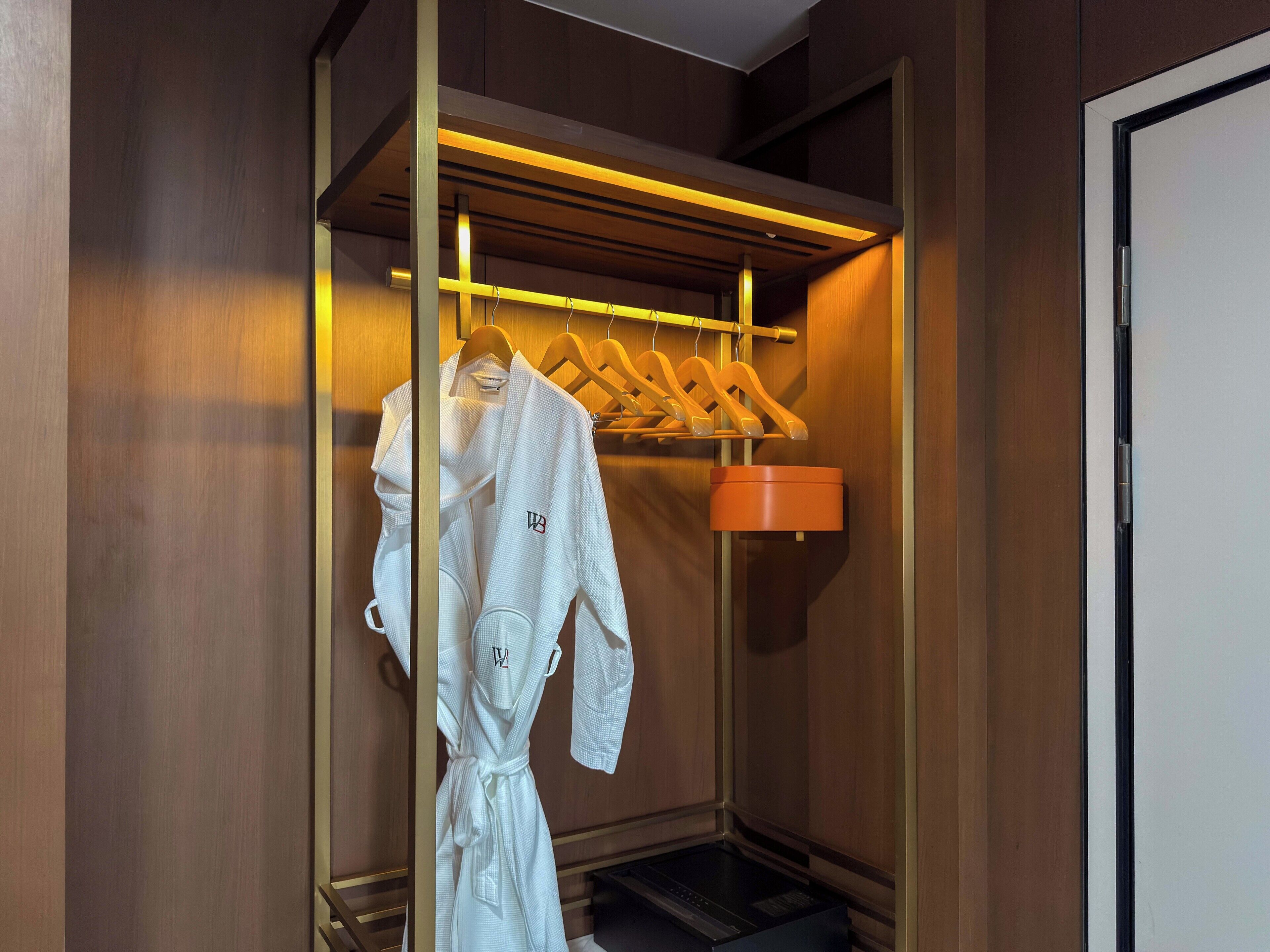 Ein begehbarer Kleiderschrank mit Holzvertäfelung, ein Bademantel an einem hölzernen Kleiderbügel und eine kleine, orangefarbene Aufbewahrungsbox.