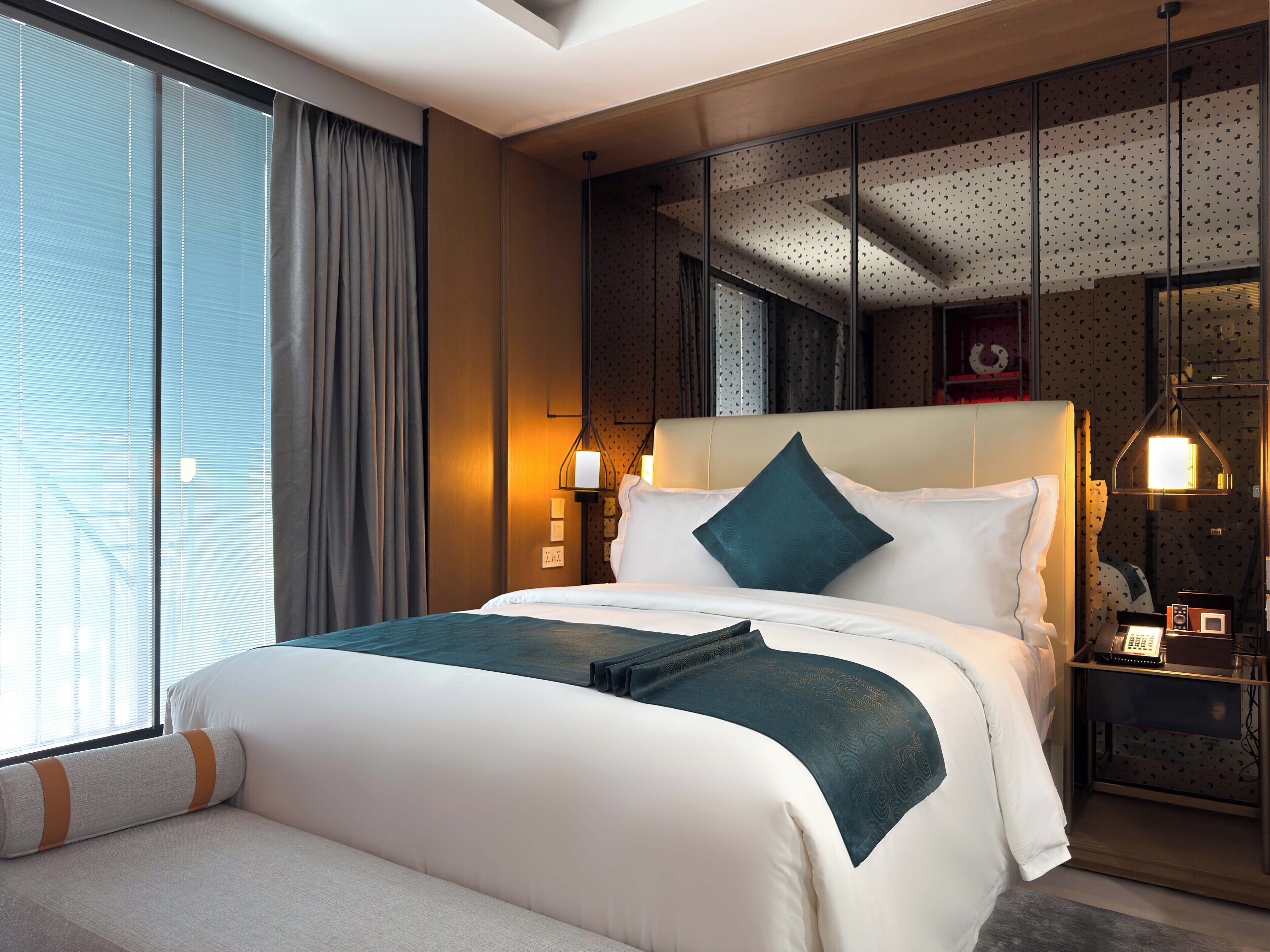 A modern hotel room with a large bed, a bedside table with a lamp, and a view of the city through large windows.