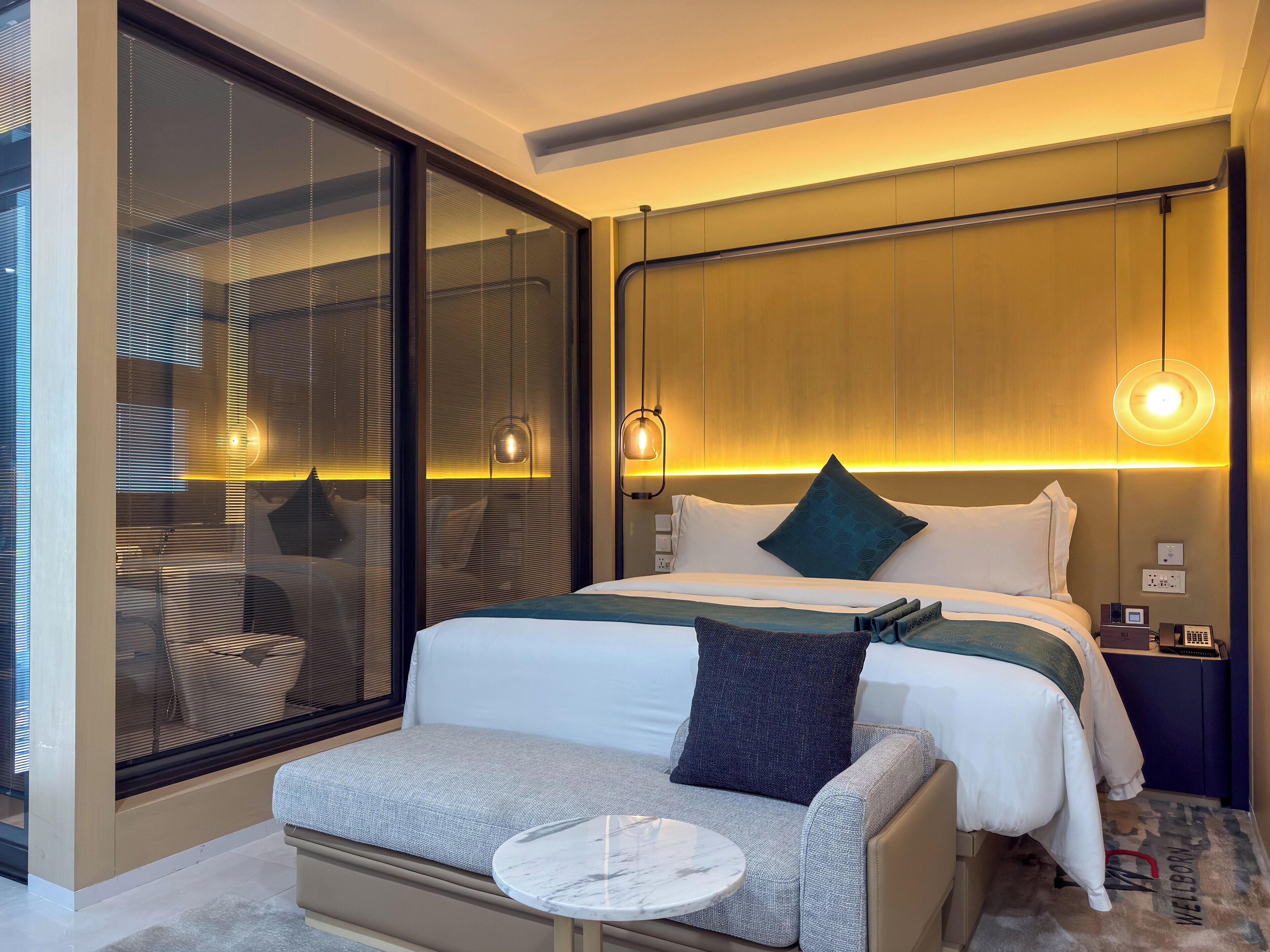 A modern hotel room with a large bed, a sofa, a small round table, and a large window with blinds.