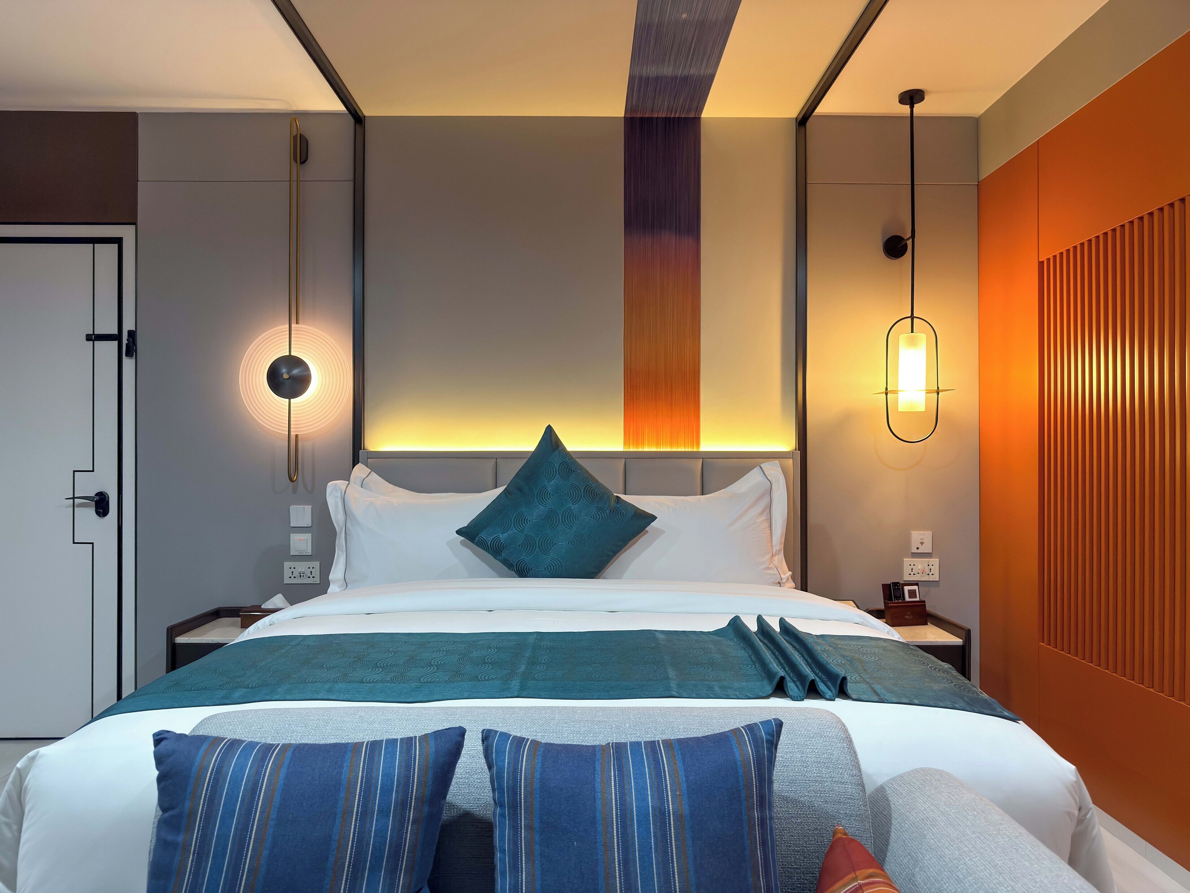 A modern hotel room with a neatly made bed, a headboard with a vertical light panel, and a wall-mounted light fixture.