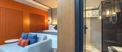 A modern hotel room with a sofa, a bed, a small table, and a bathroom with a toilet and shower.