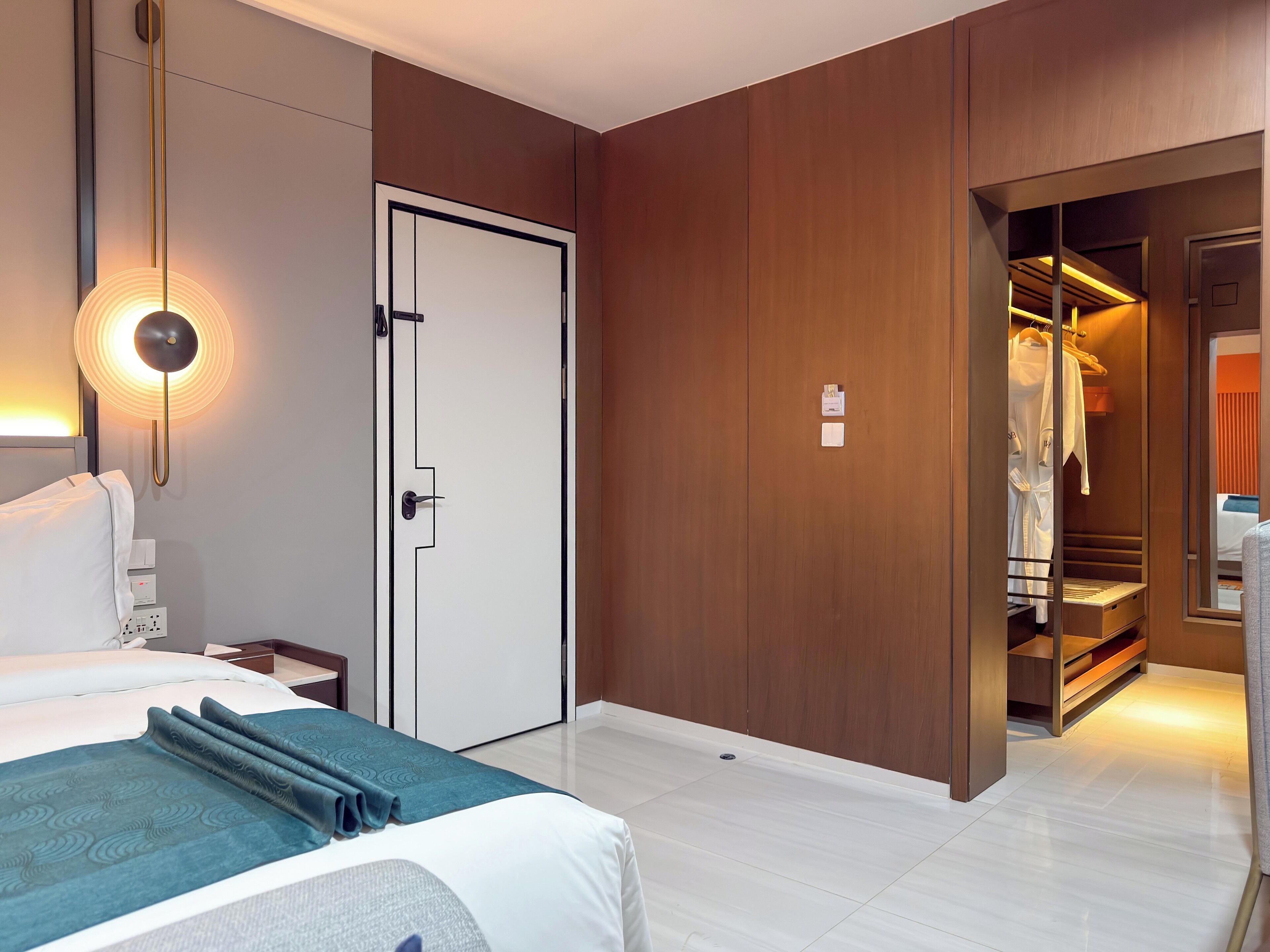 A hotel room with a bed, bedside table, lamp, wardrobe, and a mirror reflecting the room.