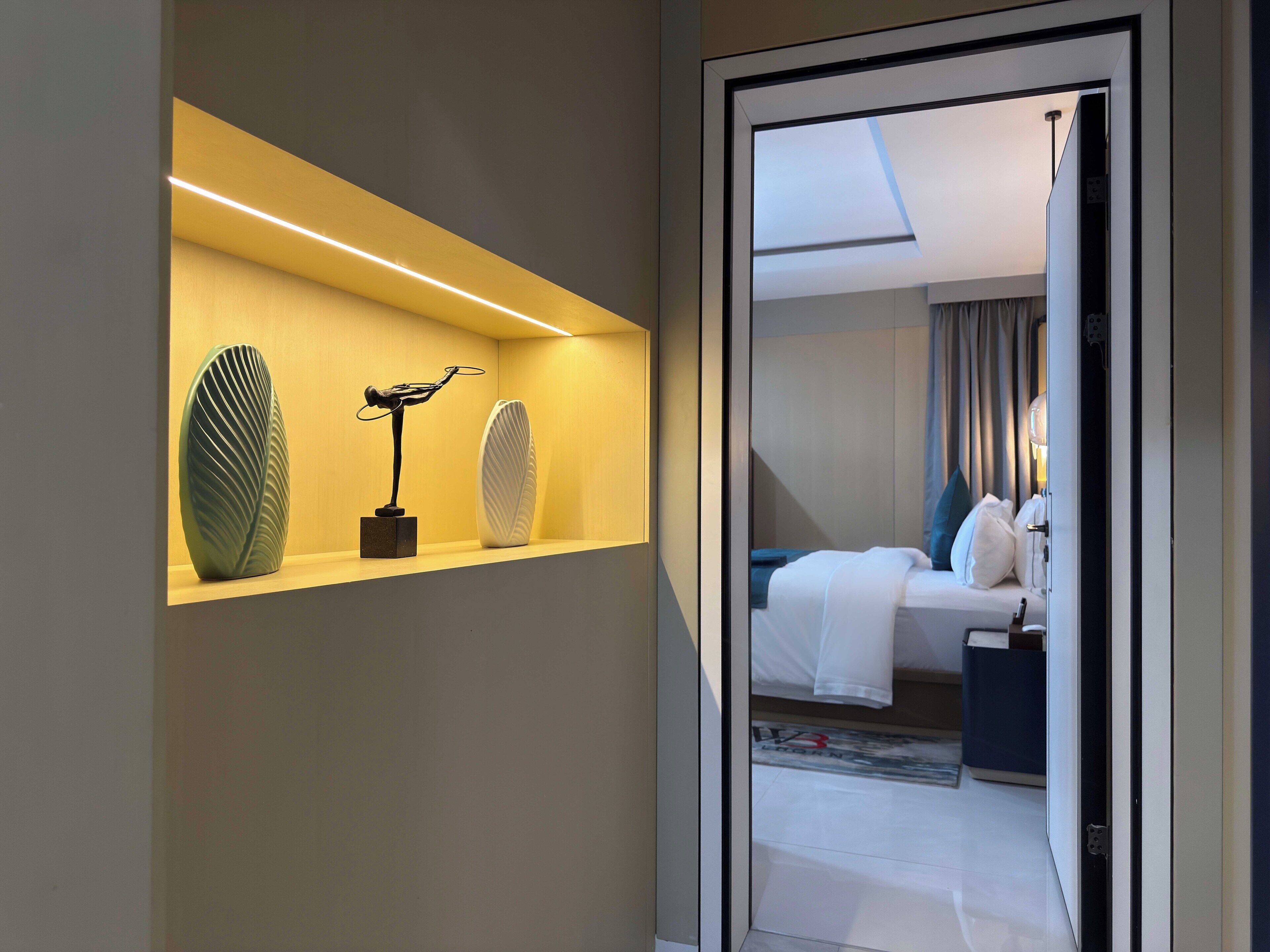 A hotel room with a bed, a nightstand, and a bedside table. There is a window with curtains, and a hallway leading to the room.