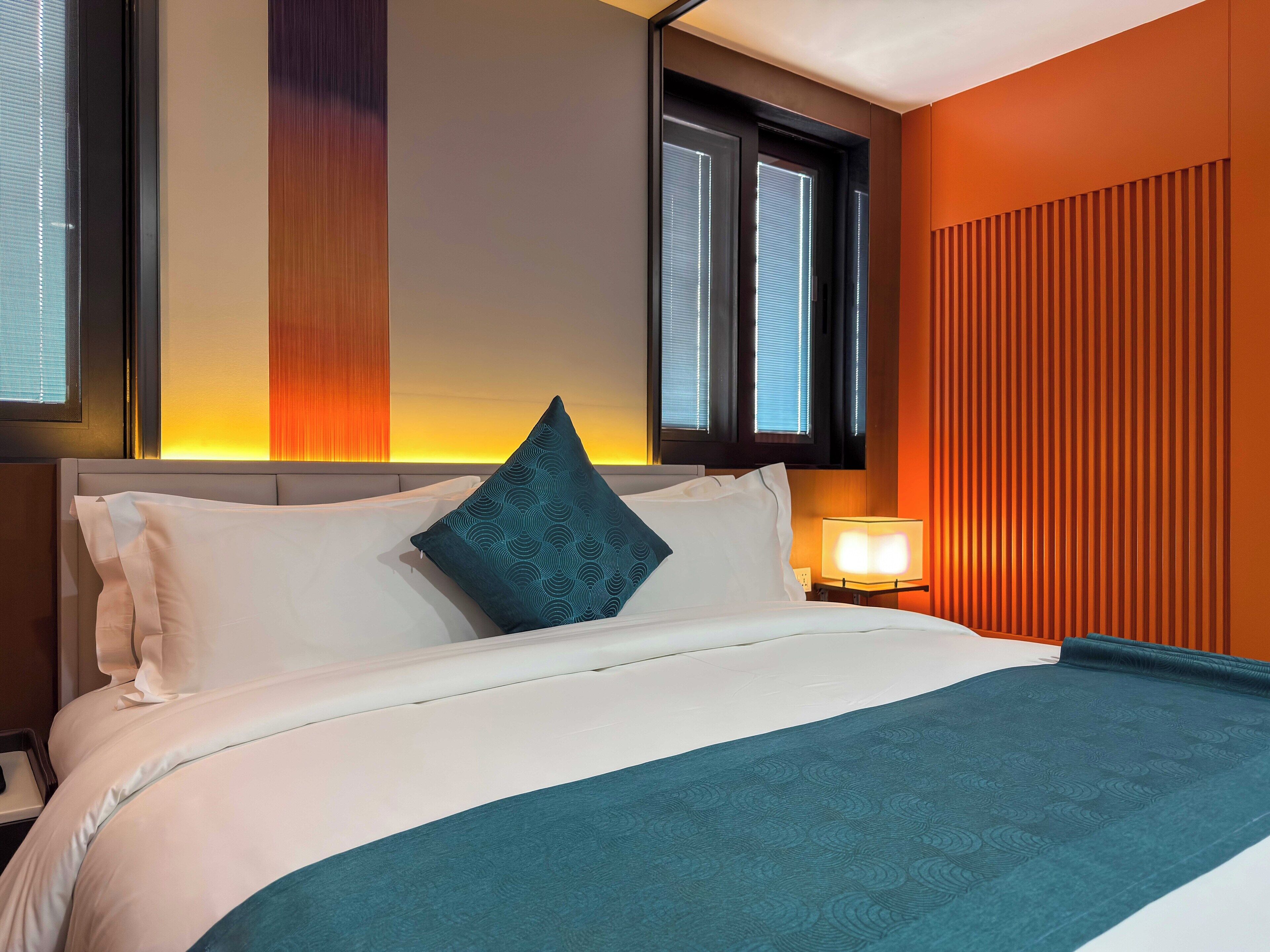 Una habitación de hotel con cama, almohadas, mesita de noche, lámpara y ventana con persianas.