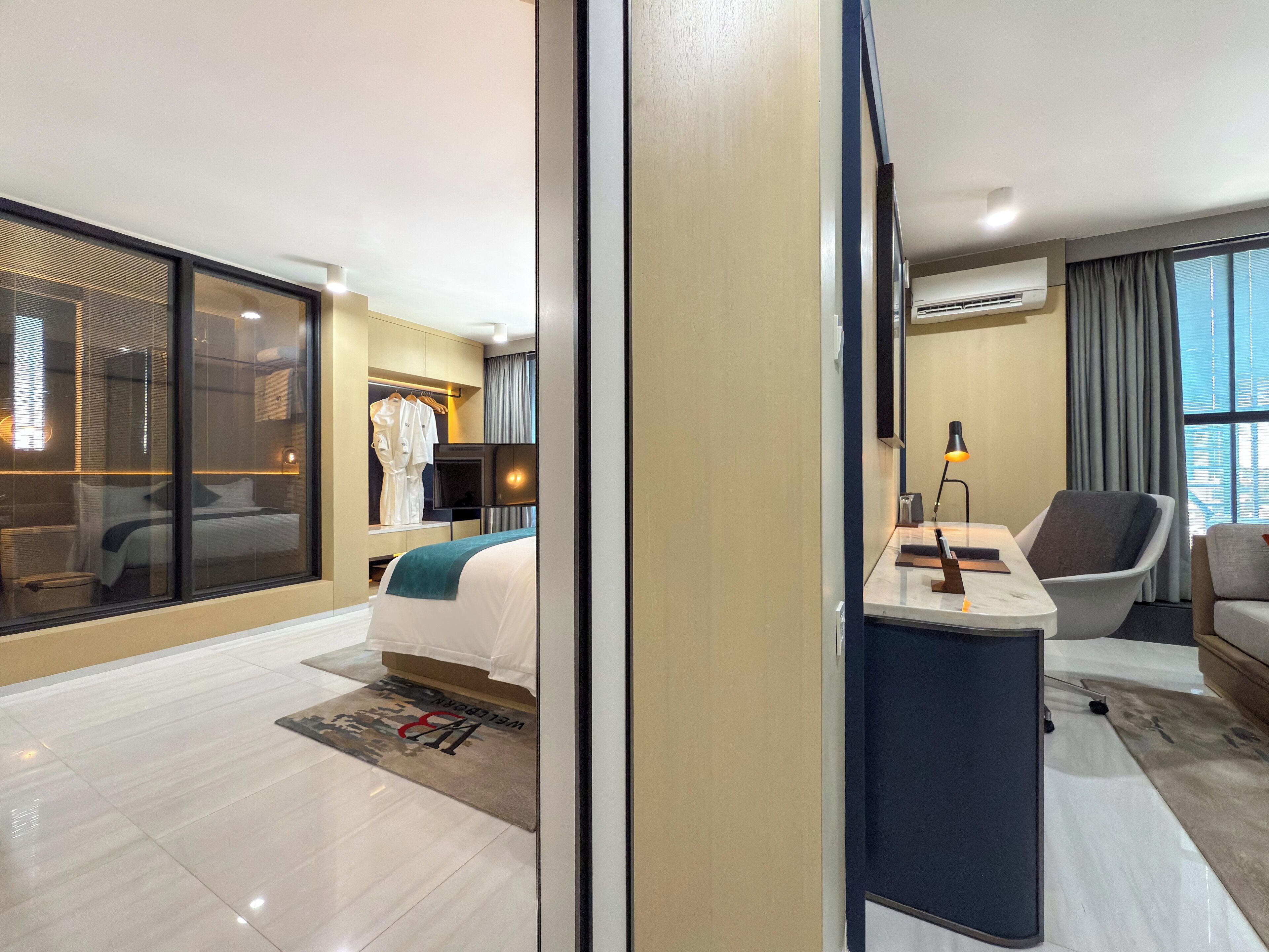 A modern hotel room with a large bed, a desk with a chair, a wardrobe, and a large window with curtains.