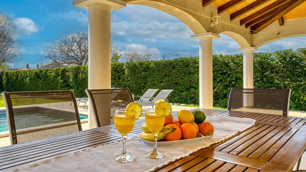 Un espace de restauration en plein air avec vue sur la piscine, comprenant une table avec un panier de fruits et des boissons, ainsi que des chaises longues.