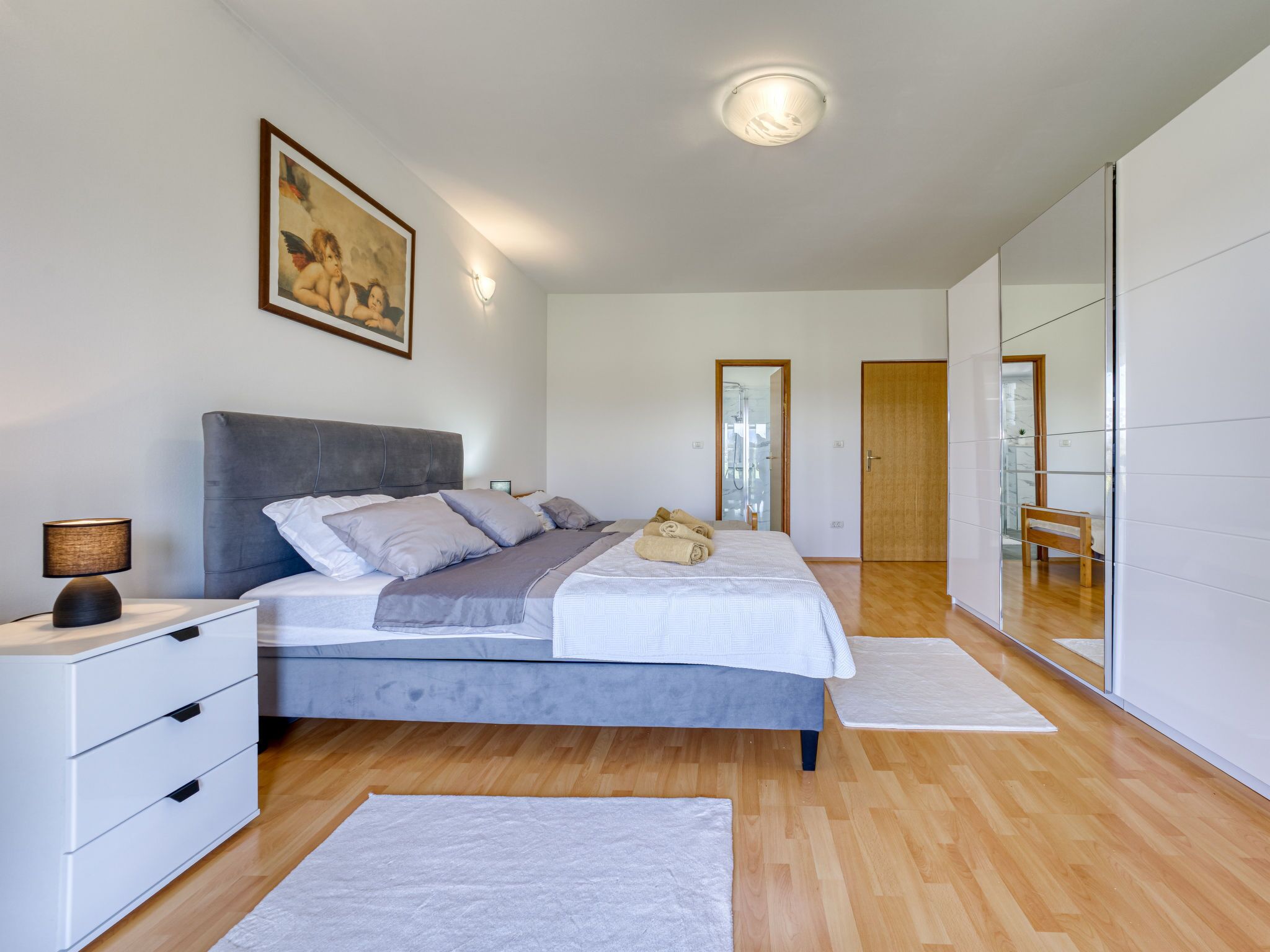 Een moderne slaapkamer met een groot bed, nachtkastjes, een lampje, een schilderij, plafondverlichting, een spiegel en een houten vloer.