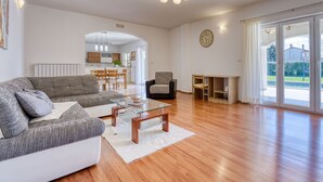 Un salon spacieux avec un canapé, une table basse et une cheminée. On aperçoit une cuisine au fond et une grande fenêtre donnant sur l’extérieur.