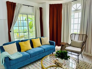 A living room with a blue sofa, a wicker chair, a glass coffee table with a plant, and a view of greenery outside. - Falcon CoralReef-Gated-24/7 Security-Airport Transport (Montego Bay)