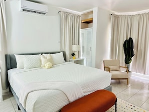 A bedroom with a large bed, a bedside table with a lamp, a chair, and a potted plant.