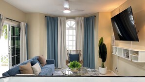 A modern living room with a sofa, a round glass coffee table, a chair, a TV mounted on the wall, and a large window with curtains.