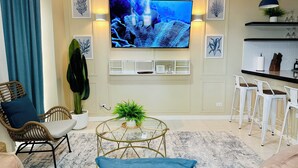 A living room with a flat-screen TV, a coffee table, a sofa, a chair, and a small kitchen area with bar stools.