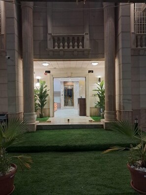Une entrée d’immeuble avec un chemin central bordé de colonnes, menant à une porte en verre.
