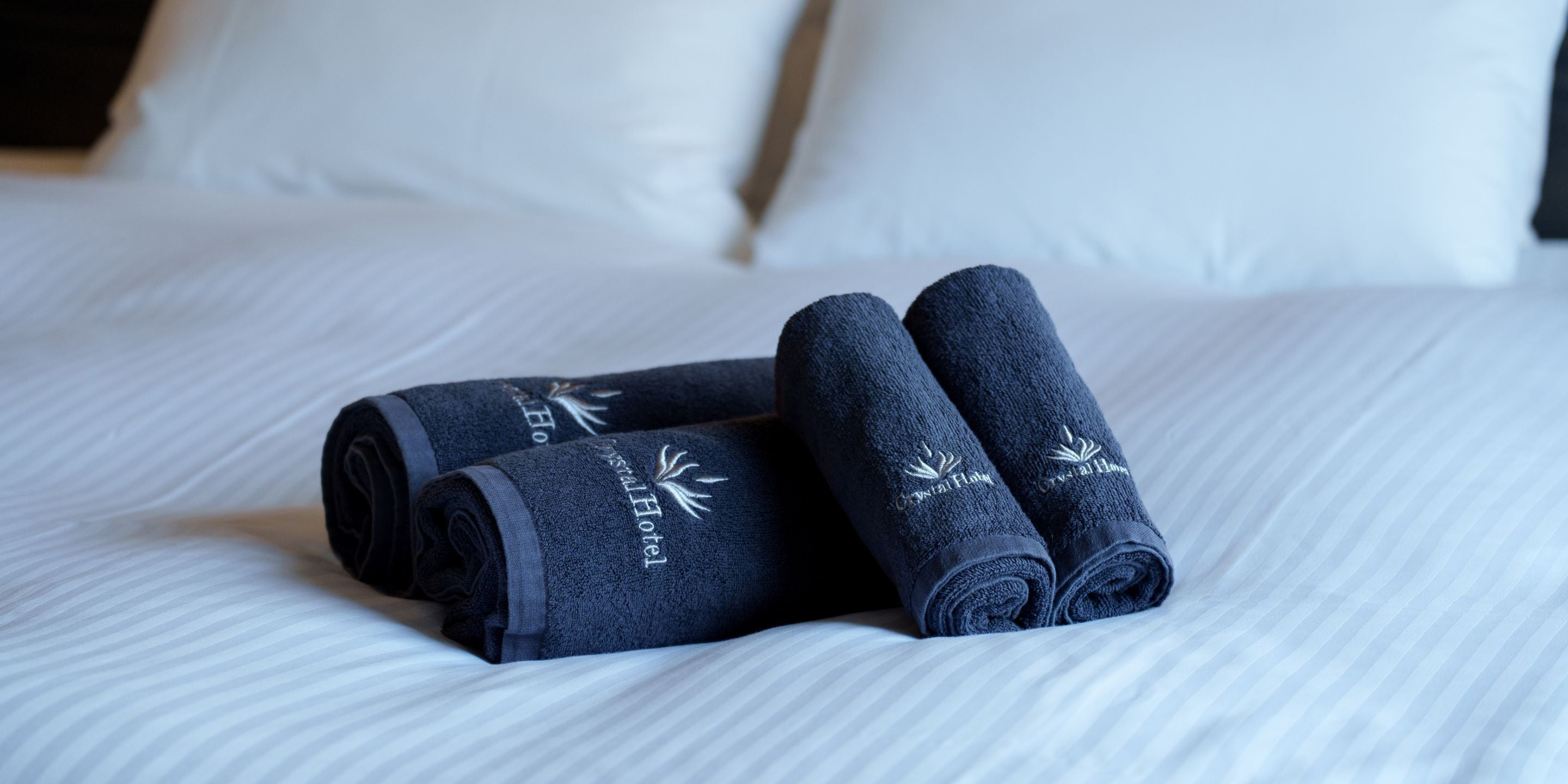 A neatly made bed with white linens and two rolled towels with embroidered text.