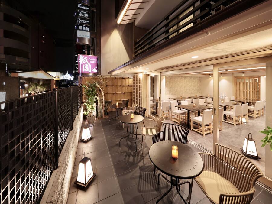 Un área de comedor al aire libre con mesas y sillas, una cerca de madera y un edificio con señalización al fondo.