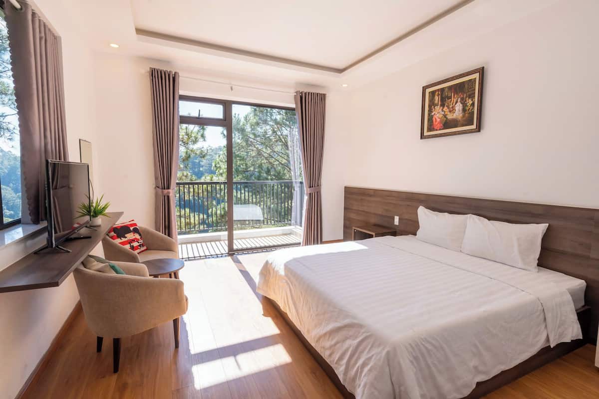A hotel room with a large bed, a TV, a desk with a chair, and a balcony with curtains.