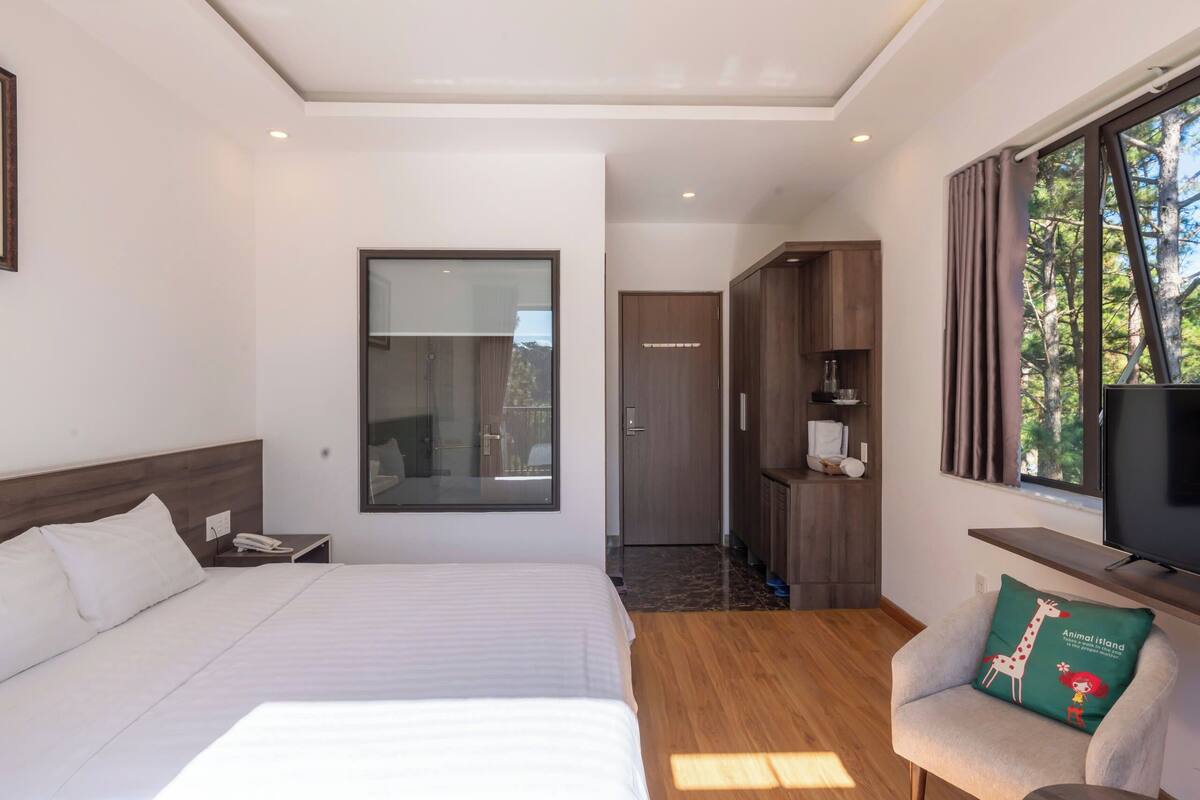 A modern hotel room with a large bed, a TV, a sofa with a giraffe pillow, and a view of trees through the window.