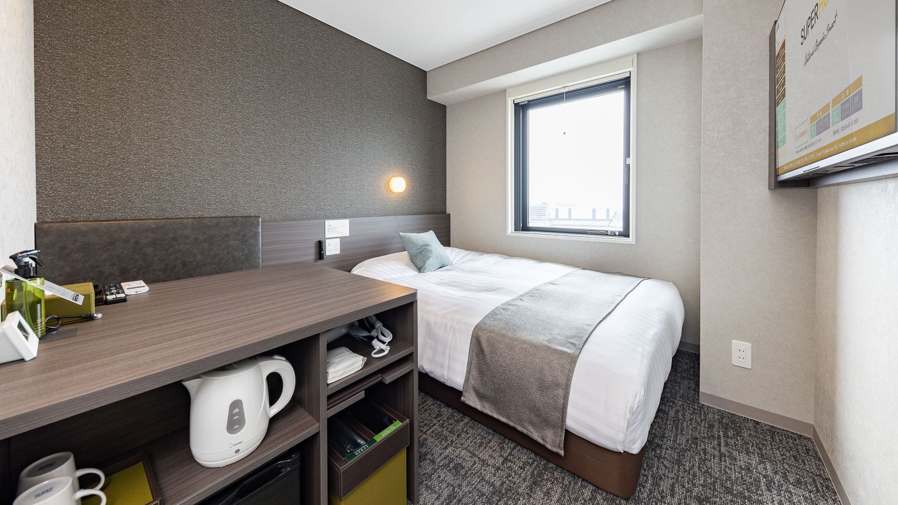 Une chambre d’hôtel avec un lit, un bureau doté d’une bouilloire, une fenêtre et un lampadaire fixé au mur.