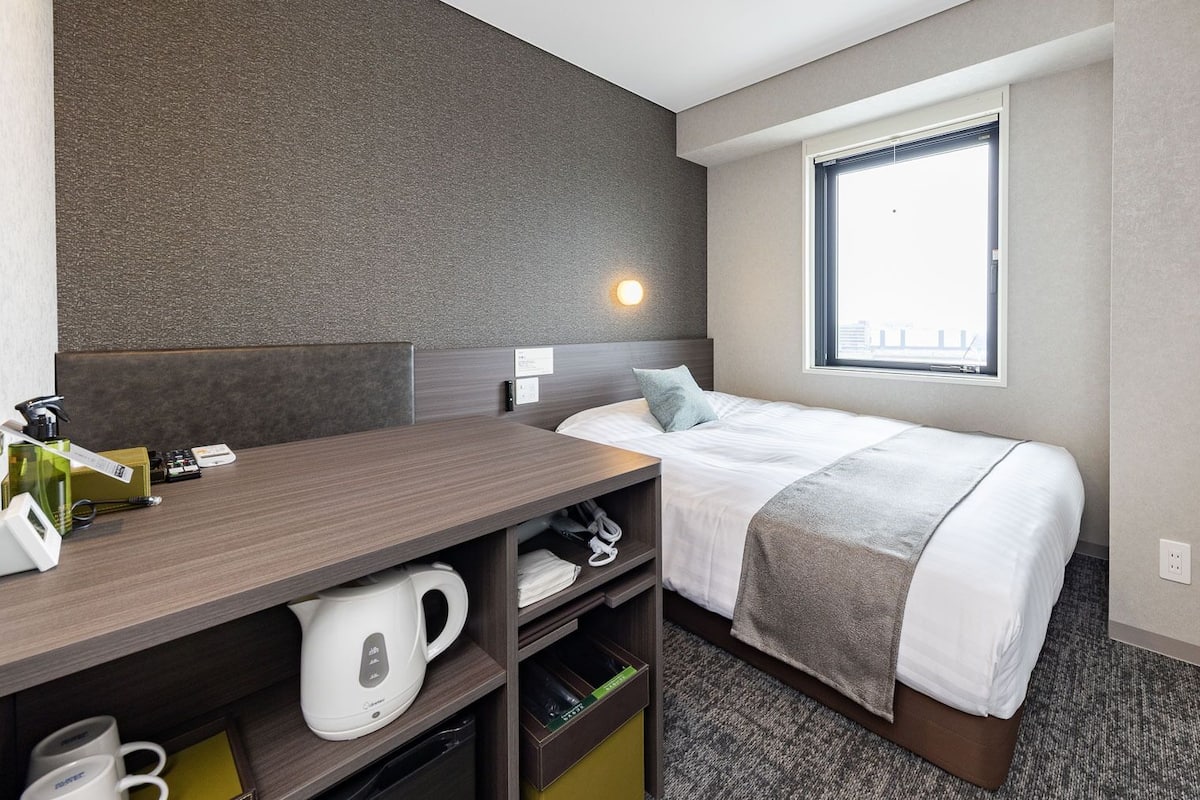 Ein Hotelzimmer mit einem Bett, einem Schreibtisch mit Wasserkocher, einem Fenster und einer Wandleuchte.